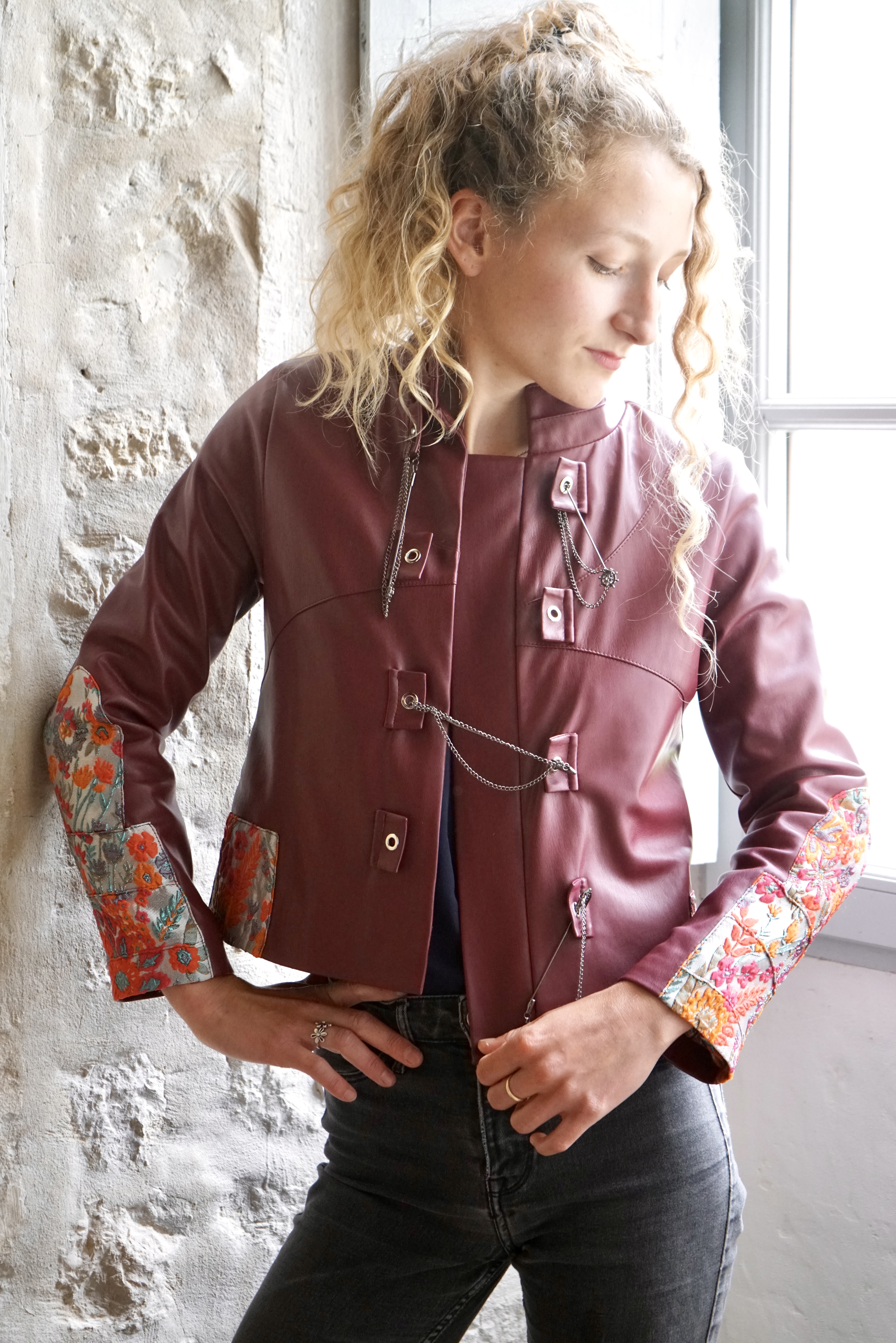 Modèle portant une veste en cuir végétalien sur mesure en rouge-brun foncé avec des patchs floraux orange-rouge brodés, un design inspiré du guerrier mongol, des fermoirs métalliques uniques, une doublure luxueuse assortie aux patchs brodés extérieurs